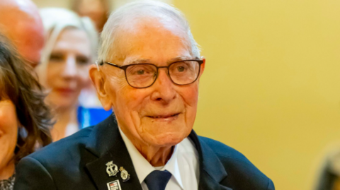 A man with grey hair and dark framed glasses in a navy jacket with metallic pins on the lapel and a white shirt with a dark tie