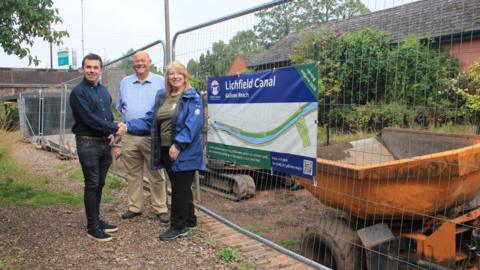 Three people are stood next to a poster on railing with machinery behind it. A man in jeans and a navy blue shirt is shaking hands with a woman wearing a green t-shirt and blue coat. The third person is a man with a light blue shirt and cream-coloured trousers.