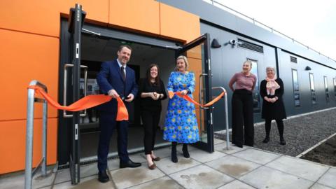 Councillor Gary Allen with short brown hair, blue suit, white shirt and maroon tie holding a cut orange ribbon with volunteer Natalie Whittaker who has long brown hair, black top, black trousers, brown shoes and is holding a pair of scissors. Councillor Pamela Hargreaves has short blonde hair with a blue patterned dress, black boots and is holding a cut orange ribbon. Leader of Hartlepool Borough Council, Danielle Connolly has blonde hair tied back with a peach coloured top and black trousers. Start Head of Service, Sally Harris and Clinical Lead has short grey hair, glasses, a black dress, black leggings and black shoes. They are stood outside the entrance to an orange and dark grey cladded building officially opening it by cutting the ribbon.