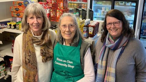 Three female volunteers stand in the shop smiling into the camera. They stand with food products on shelves around them.  The middle volunteer wears a green Aston on Clun Community Shop apron.