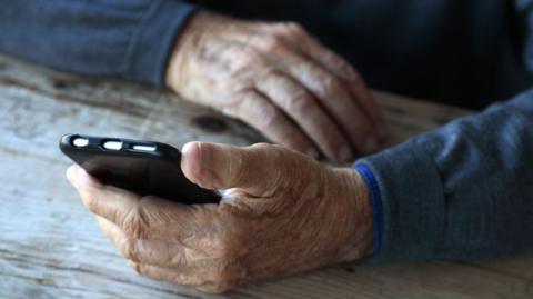 An elderly person holding a mobile phone. We can only see their hands which hold a black, rectangular-shared mobile. Their wrists are also visible, resting on a wooden table and they have light blue sleeves.