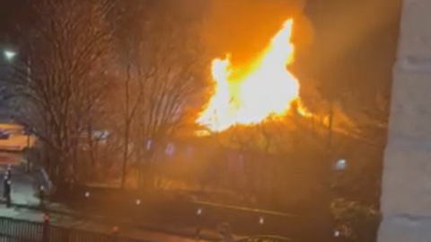 Large flames can be seen coming through the roof of the building on Canal Street. Bare trees are silhouetted in the street in front of the fire. 