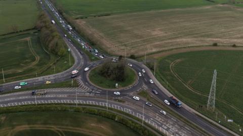 An aerial view of a large roundabout with a grassy central island and three exits. There are trees in the centre and multiple lanes coming off at each exit. The roundabout is set between grassy fields. Buildings of a residential area are int eh background.