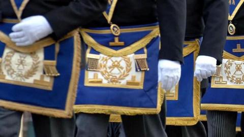 Masons march in a procession. Pictured are men from waist height wearing white gloves and wearing the Freemasons coat of arms embraided with gold thread on a royal blue background