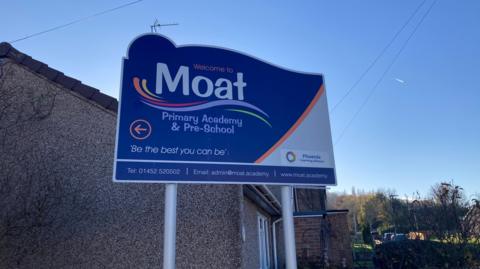 A large blue metal sign says Welcome to Moat Primary Academy, with the school motto "Be the best you can be" written on the lower section of the sign 