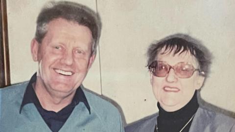 Two people in an old photo - on the left is a man with brown hair in a blue jumper and black shirt. On the right is Patricia, a woman with a black roll neck, a grey blazer and glasses.