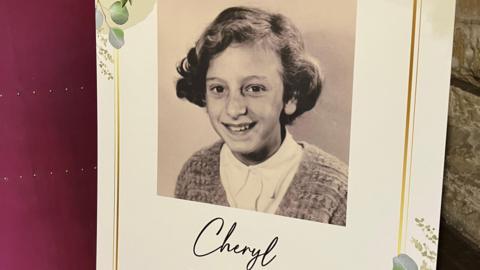 A black and white photo of a young girl smiling with a bob haircut and a knitted cardigan. The photo is on a poster propped up against a wall. It says Cheryl underneath and there is a gold frame printed around the photo.