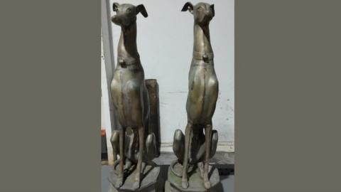 Two dog statues depicting slim large dogs sitting upright on plinths - they have identical collars.