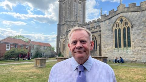 Man in a shirt and tie looking at the camera. He's stood in a churchyard with a historic church behind.