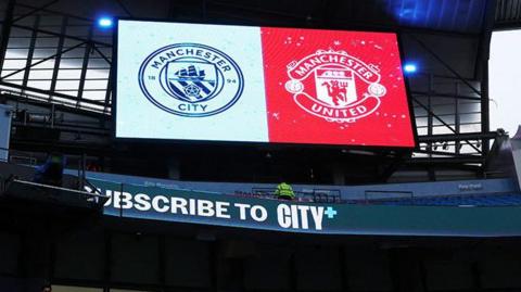 Inside the Etihad Stadium before the Premier League match between Manchester City and Manchester United. Both clubs' emblems are shown side by side on the big scoreboard screen.