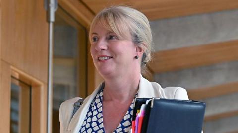 Shona Robison is walking down a wood-panelled corridor in the Scottish Parliament holding a black leather-bound folder. The head-and-shoulders shot shows Robison with her blonde hair tied back and wearing a blue and white top with a white blazer.