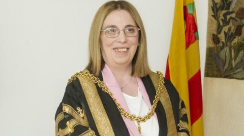 Mayor Jeanette Whalen wearing the existing robe at her mayor-making earlier this year. She has long blonde hair and is wearing glasses. She has a gold chain around her neck and is wearing a black robe decorated with gold embroidery