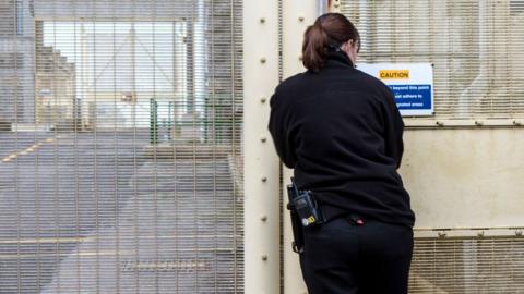 A female prison officer dressed in black with brown hair tied into a ponytail unlocks a prison gate at HMP/YOI Portland, Dorset.