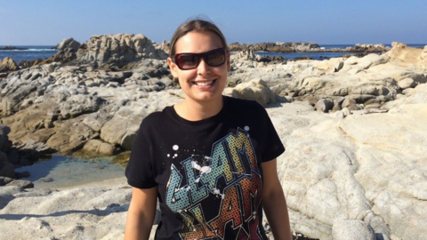 A woman is smiling, wearing sunglasses and a black graphic t-shirt, appears to be standing on some rocks at a seaside.