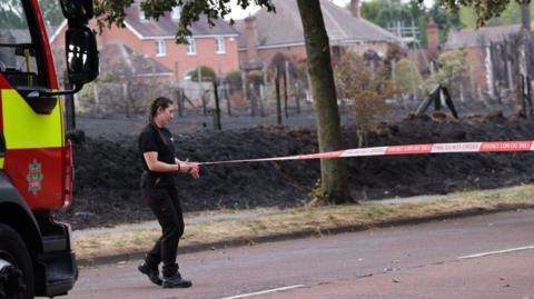 A fire fighter with her hair in long plaits pulls a red and white do not cross tape across a road. Behind her is blackened grassland with houses in the distance.