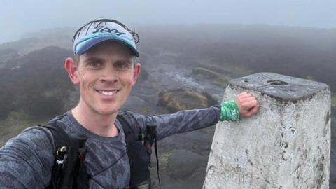 Fell runner Ben Morris takes a selfie at a summit trig point in the countryside. He is wearing a grey top and a black trail running vest and a hat and rests his hand on the trig point. The peaks in the background are covered in mist.