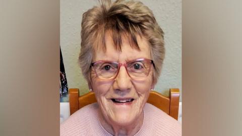 Jean Harris, wearing glasses and a pink sweater, smiling