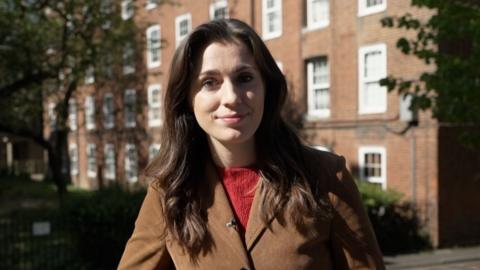 A woman stand in front of housing with the sun shining on her face.