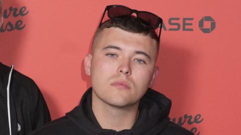 Close shot of a dark haired young man with short brown hair, a black hooded jacket and dark sunglasses on top his head