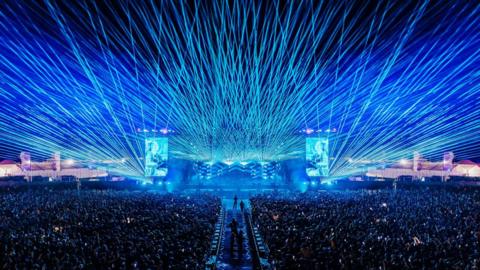 Hundreds of blue laser beams shine out from the main stage at Boardmasters during a set at the 2025 festival. Thousands of people are stood in front of the stage watching the show.