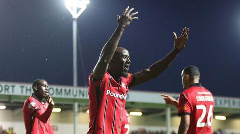 Albert Adomah has his arms up in the air celebrating a recent goal, wearing his Walsall home red and black kit