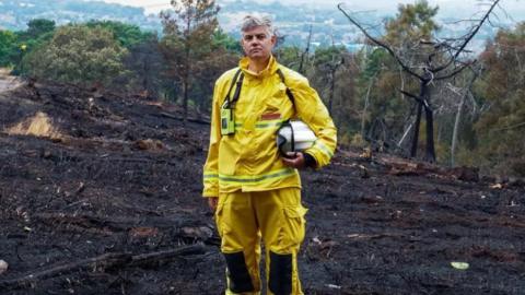 The picture shows fire chief and wildfire advisor Dave Swallow standing in burnt countryside after a moorland fire near Birmingham in 2022. He is wearing yellow firefighting gear and has a helmet, with equipment round his neck. The ground is charred and black.