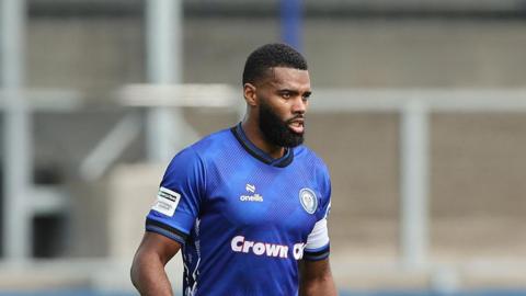 Ethan Ebanks-Landell playing for Rochdale on a sunny day, wearing a captain's armband on his left arm, about to play a pass 