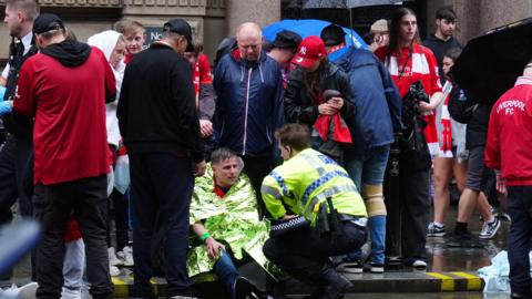 Police officer crouched down talking to man wrapped in emergency service blanket at he sits on the floor. Lots of fans are standing behind them in red, some are wearing coats or holding umbrellas to protect them from the rain