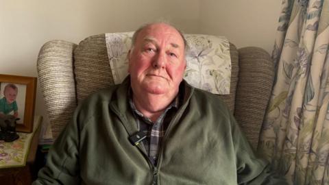 A man with grey hair sits in an arm chair wearing a checked shirt and a green zip up fleece. The arm chair is striped beige, with a patterned head rest that matches the curtain on the right of the picture. To the left, there is a picture of a young child on a side table.