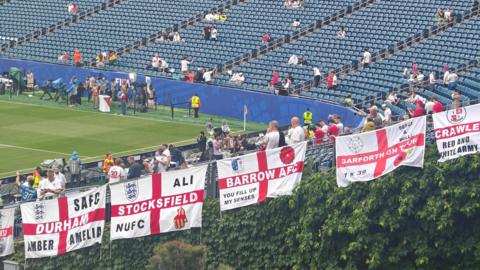 A string of England flags attached to the side of a rail by the stands at the Boston stadium where England played against Ghana. The flags all feature the names of different towns and local clubs. It includes one with the words Barrow AFC across the horizontal line of the red cross. In the top left corner it has the Barrow AFC badge, the top right has a red rose, the bottom left has the phrase "you fill up my senses" and the remaining quarter is blank.