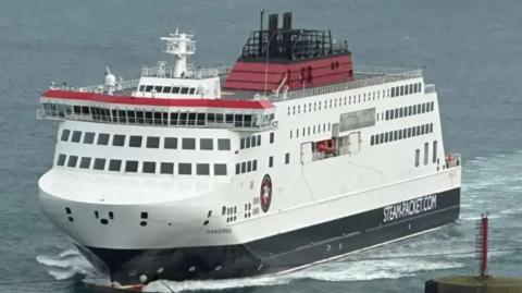 The Manxman sailing towards Douglas in calm waters. It is painted in the Steam Packet's livery of black, white and red and has the company's logo on the side at the front and the words STEAM.PACKET.COM further towards the back.