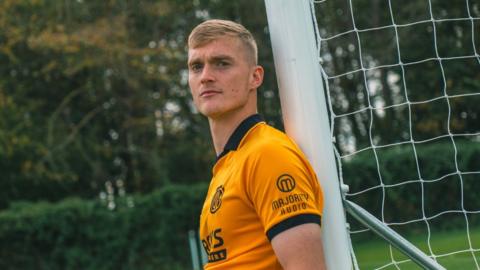 Cathal Heffernan, in the colours of Cambridge United, leaning his back against the goalpost while looking towards the camera