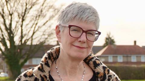 A woman with short cropped grey hair is wearing glasses and a leopard print coat. She is standing outside and smiling