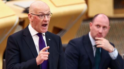 John Swinney has his hands out in front of him as he speaks in the Holyrood chamber. He has a bald head and is wearing glasses. He is wearing a dark suit, white shirt and purple tie