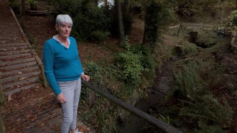 A woman with short, white hair, a blue jumper and grey trousers standing on a small wooden bridge above a small river.