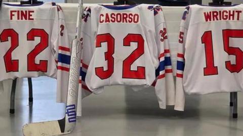 An image showing the hockey jerseys of the three boys who were killed draped over a long table. The jerseys are white with red names and numbers, and with a navy blue collar and stripe on each sleeve. Fine wears a 22 jersey. Casorso wears a 32 jersey and Wright wears a 15 jersey. Beside each jersey is a hockey stick. 