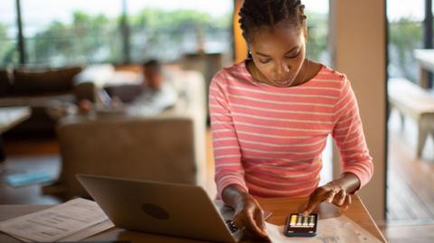 A woman sits at her kitchen table using the calculator on her phone. Her laptop and some bills are also on the table. She is wearing a pink and white striped top.