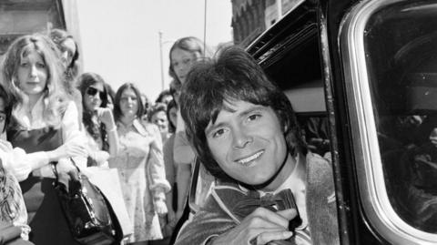 A black and white image of Sir Cliff Richard leaning out of a Mini. He is wearing a dinner suit and bow tie. There is a crowd of women behind the vehicle looking towards the camera.