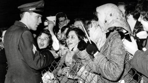Elvis greeting fans at Prestwick Airport in 1960. He is dressed in his military uniform. The fans are mostly female and looking at him with excitement and adoration. 
