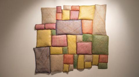 The image shows a textile-based wall artwork composed of many small, padded rectangular sections arranged into an irregular grid-like shape. The piece is mounted against a plain, light-coloured wall.