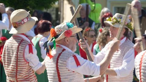 Three Morris dancers in Morris dancing costumes, waving wooden sticks. There is a crowd of people in the background.