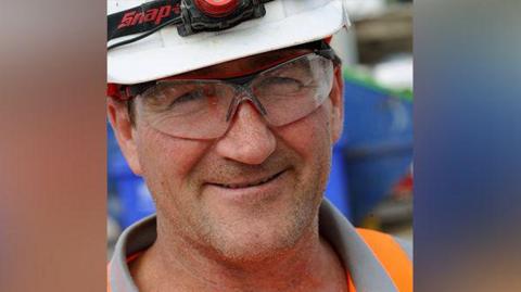 Frank Hooper close up of face wearing hard hat, safety specs and organge hi vis vest.