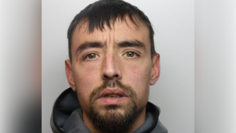 A mugshot of a man with short black hair, a beard and grey hooded top.