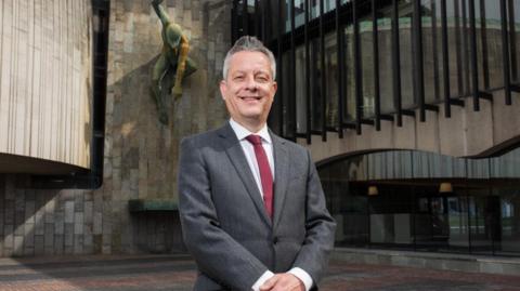 Library image of Councillor Nick Kemp standing outside Newcastle Civic Centre. He has grey hair, brushed back, and is wearing a grey suit, white shirt and maroon tie.