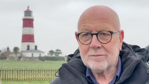 A head and shoulders image of Harry Blathwayt. He is wearing a dark, hooded top and glasses. Happisburgh Lighthouse is fully in view over his right shoulder. It has three red and two white stripes around it, below the lantern, and a cottage at its base.