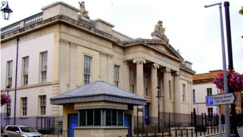 Shows a courthouse with four pillars at the entrance and a security sangar to the left. A silver-coloured car is on the left of the picture and, on the right, a signpost pointing to Bishop Street car park and saying it has 130 spaces.