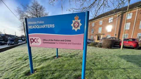 A three-legged sign reading Wiltshire Police headquarters on grass outside a building
