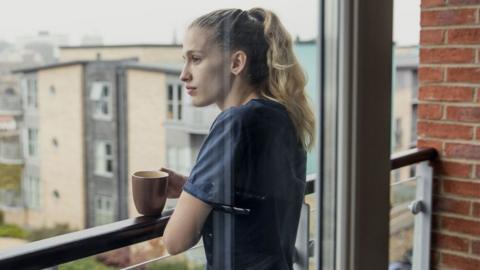 A woman with blond hair in a ponytail and a blue t-shirt stands on her balcony with a cup of tea and looks out wistfully at nearby residential buildings