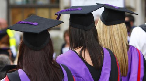 A file photo of university graduates. They are standing with their back to the camera and are wearing black robes with purple hoods and black graduation caps. All three have long straight hair.