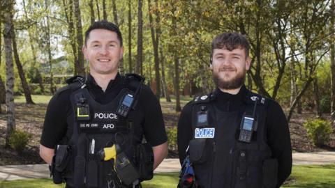 PC Sam Clover and PC Jacob Edwards standing outside next to each other. They are both wearing black police uniform and are looking directly at the camera and smiling. There are trees behind.